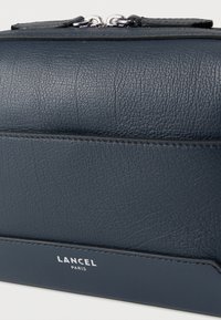 Σκούρο μπλε δερμάτινο πορτοφόλι με υφή, που διαθέτει λείο στοιχείο και λεπτομέρεια με λογότυπο. Διαθέτει κλείσιμο με φερμουάρ και συνδεδεμένο μεταλλικό εξάρτημα.