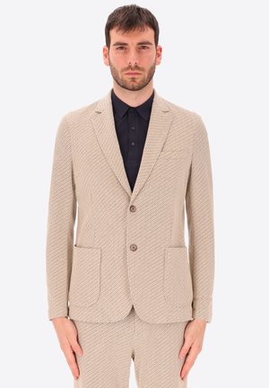 Uomo che indossa completo beige strutturato con blazer a due bottoni e pantaloni coordinati, abbinato a camicia nera abbottonata, in piedi davanti a sfondo uniforme.