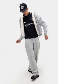 Calças de fato de treino cinzentas, uma t-shirt preta de manga curta com logo, e um casaco cinzento com fecho de correr usado aberto. Boné e sapatos pretos completam o conjunto.