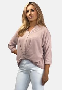 Jasnoróżowa, luźno dopasowana bluzka z wyciętym kołnierzykiem i rękawami długości trzy czwarte, w połączeniu z białymi, dopasowanymi spodniami. Gładka tekstura.