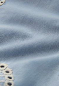 Tissu bleu clair doux avec une texture subtile et de petits détails floraux découpés brodés en crème le long du bord.