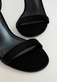 Sorte højhælede sandaler med en glat læderfinish og et bredt velourbånd over tåen. Sålen er rundet og tekstureret.