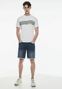 Bílý tričko s horizontálními černými pruhy a textem, doplněné o ohrnuté tmavé riflové šortky a bílé tenisky. Jednoduchý, neformální design.