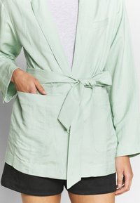 Robe en lin vert clair avec une ceinture à la taille, des manches amples et deux poches avant, portée par-dessus une chemise grise et un short foncé.
