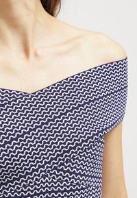 Off-the-shoulder toppi tummansinisellä värillä, jossa on valkoinen zigzag-kuvio. Sisältää sileän tekstuurin ja istuvan muotoilun kaula-aukolla.