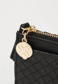 Pochette en cuir noir texturé avec une fermeture éclair en ton doré et un charm décoratif en or avec une gravure florale.