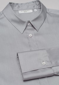 Grå fitted skjorte lavet af glat stof, med spredt krave, hvide knapper og en enkelt manchet med knaplukning.