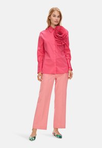 Pink collared shirt with a large pleated floral accent on one shoulder, paired with wide-legged striped pink pants and floral flats.