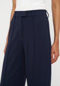 Femme portant un pantalon large taille haute bleu marine et un top blanc sans manches rentré, debout avec un bras légèrement plié.