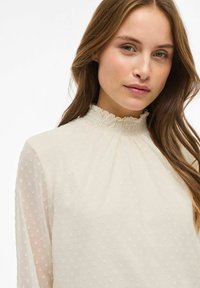 Hvid bluse med prikker, høj rynket halsudskæring; gennemsigtige lange ærmer; blød tekstur; detaljer med subtile mønstre på stoffet.