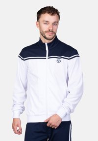 Witte zip-up jas met marineblauwe kraag en strepen. Bevat een logo op de borst. Soepele stof met elastische manchetten en zoom. Subtiel ontwerp.