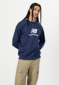 Tumši zils džemperis ar garām piedurknēm, ar baltu "new balance" logo. Kokvilnas maisījuma audums, ribotas aproces, brīva piegriezuma, valkāts ar bežām cargo biksēm.