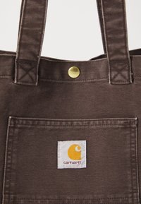 Brown canvas tote bag featuring a square patch with a yellow logo and brand name. Includes a gold snap button and sturdy straps.