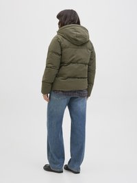 Veste matelassée verte avec capuche, au design épais, associée à un jean bleu à coupe décontractée et des chaussures noires sur un fond uni.