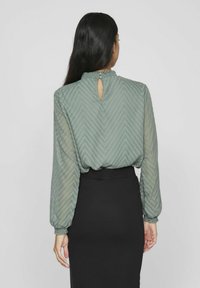 Zielona bluzka w zygzakowaty wzór z wysoką szyją i wycięciem z tyłu, zestawiona z czarną spódnicą. Bluzka ma długie rękawy i elastyczne mankiety.