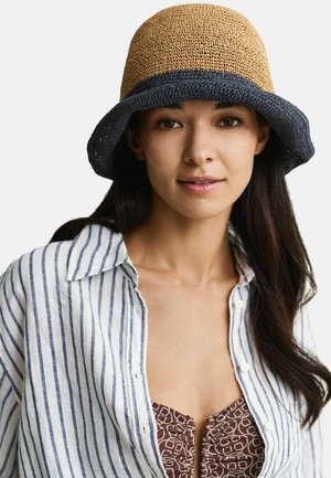 Donna con capelli lunghi e scuri che indossa un cappello a secchiello intrecciato beige e nero, una maglietta a righe bianche e blu e una maglia marrone con fantasia sotto.