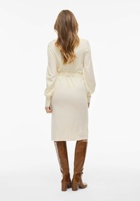 Robe tricotée couleur crème avec de longues manches ballons, ceinturée à la taille, de longueur midi, assortie à des bottes marron jusqu'aux genoux.