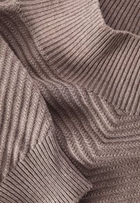 Gros plan sur un tissu tricoté taupe à textures côtelées et chevrons, montrant des sections pliées et superposées en fil doux et épais.