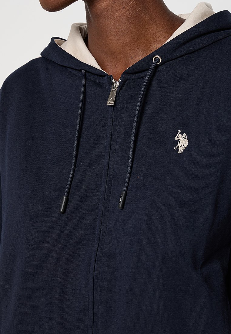 Close-up van een persoon die een marineblauwe hoodie met rits draagt, met een zilveren paard en ruiter logo op de linkerkant van de borst en een beige voering van de capuchon zichtbaar.