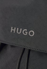 Black fabric backpack featuring a textured surface, drawstring closure, and embossed "HUGO" logo on the front flap.