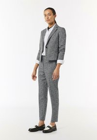 Grau strukturierter, maßgeschneiderter Anzug mit einer verkürzten Jacke, passender Hose und einem weißen Hemd mit Kragen. Schwarze und weiße Slipper komplettieren den Look.