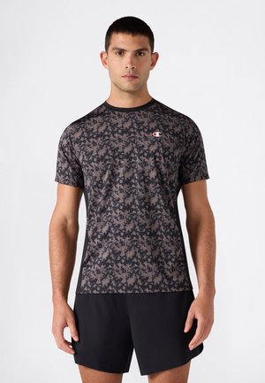 Mann trägt ein kurzärmeliges Sportshirt in schwarz und dunkelgrauer digitaler Tarnung und schlichte schwarze Shorts, steht vor weißem Hintergrund.