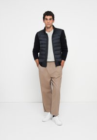 Homme portant un pantalon large beige, des baskets blanches, une chemise gris clair et une veste noir matelassée avec des manches en tricot, se tenant les mains dans les poches.
