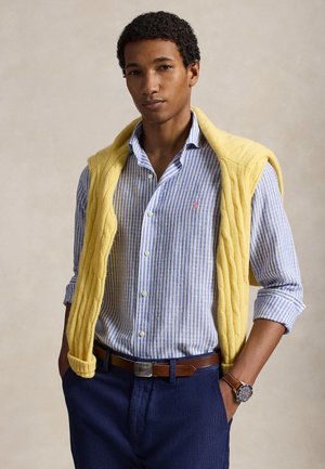 Jeune homme portant une chemise bleue à rayures, un pull jaune drapé sur les épaules, un pantalon bleu marine, une ceinture marron et une montre-bracelet, debout avec les mains dans les poches.