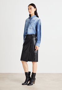 Camicia di jeans accorciata con due tasche frontali, abbinata a una gonna avvolgente in pelle nera con orlo asimmetrico e dettaglio con fibbia. Stivali neri con tacco.