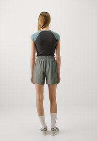 Kobieta stoi tyłem, ubrana w krótką czarną i zieloną bluzkę, zielone szorty z elastycznym pasem, białe skarpetki do kostek i szare sneakersy.