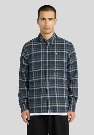 Man draagt een zwart-blauw geruit langermhemd met knopen en een klein geel logo op de borst, staand tegen een grijze achtergrond.