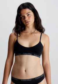 Czarny stanik braletka z regulowanymi ramiączkami, z szerokim elastycznym pasem z logo "Calvin Klein" w kontrastowym kolorze. Gładki, minimalistyczny design.