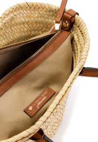 Bolso de mano de paja tejido con ribete de cuero marrón claro, cremallera abierta en la parte superior y interior de tela beige con etiqueta de cuero de la marca.