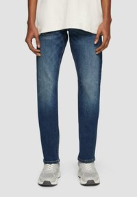 Jean en denim bleu foncé avec une coupe droite. Présente un léger dégradé, cinq poches et des coutures contrastées. Porté avec des baskets grises.