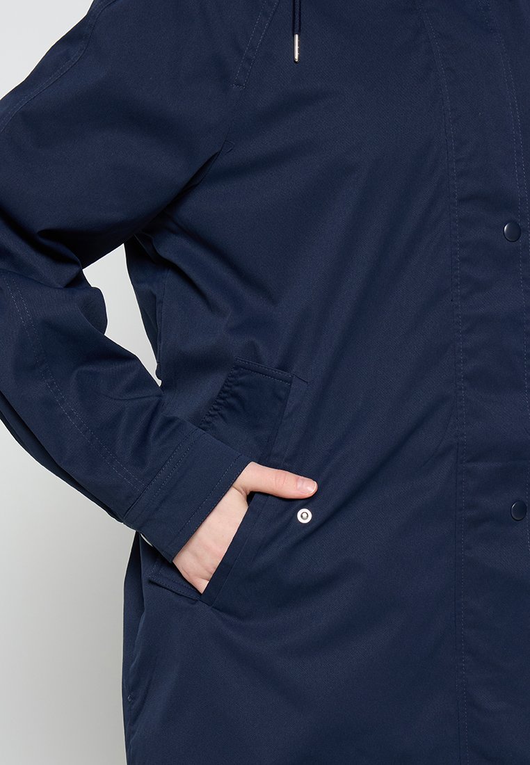 Person in einer marineblauen Jacke, mit der rechten Hand in der Seitentasche, wobei Knopf- und Kordeldetails sichtbar sind.