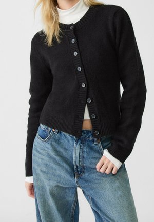 Person trägt einen schwarzen, geknöpften Cardigan über einem weißen Rollkragenpullover, kombiniert mit hoch taillierten blauen Jeans und posiert mit einer Hand in der Tasche.