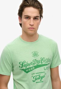 Zielona bawełniana koszulka z okrągłym dekoltem, krótkimi rękawami oraz graficznym nadrukiem w ciemniejszym odcieniu zieleni, który zawiera tekst z nazwą marki i lokalizacją.