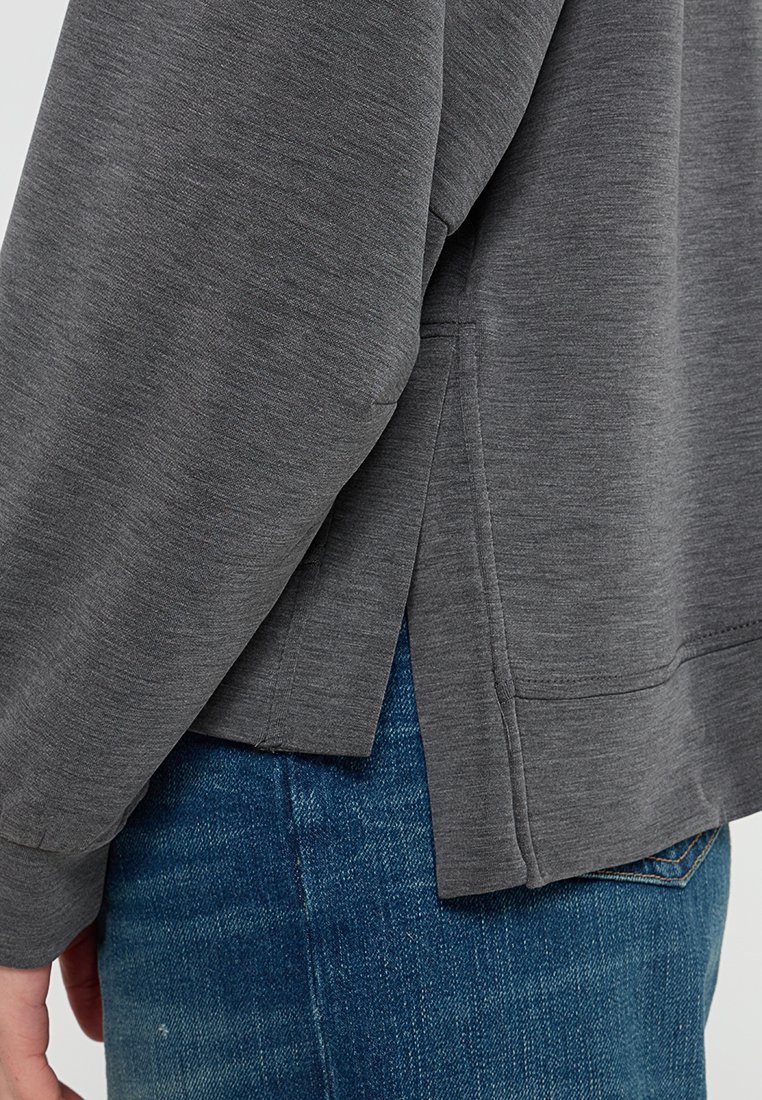 Dunkelgraues Sweatshirt mit lockerer Passform, seitlichem Schlitz, langen Ärmeln und weichem, strukturiertem Material. Kombiniert mit blauen Jeans.