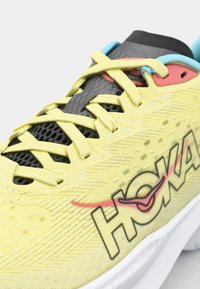 Baskets de sport jaunes avec une tige en mesh texturé, des accents noirs et turquoises, un logo HOKA bien en vue, et un design à lacets.