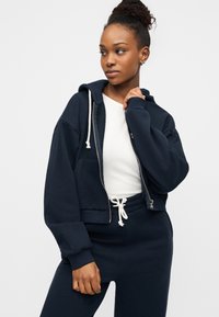 Μπλε σκούρο cropped φούτερ με κορδόνι, φερμουάρ και πλαϊνές τσέπες, σε συνδυασμό με λευκό τοπ και αντίστοιχο μπλε σκούρο φόρμας παντελόνι.