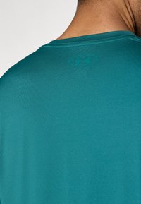 Τιρκουάζ αθλητική μπλούζα με λεία υφή και κοντά μανίκια, με λογότυπο σε λίγο πιο σκούρο τόνο στο πίσω μέρος κοντά στη λαιμόκοψη.