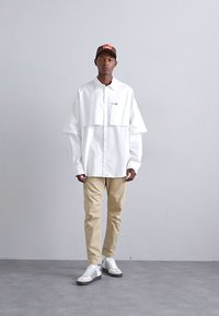Cămașă albă supradimensionată cu design în straturi, pantaloni khaki și pantofi albi cu dungi negre. Şapcă maro cu logo. Fundal gri uniform.