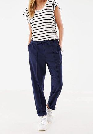 Vrouw draagt een wit en zwart gestreept shirt met korte mouwen, marineblauwe broek met trekkoord, en witte sneakers, staand met handen in de zakken.