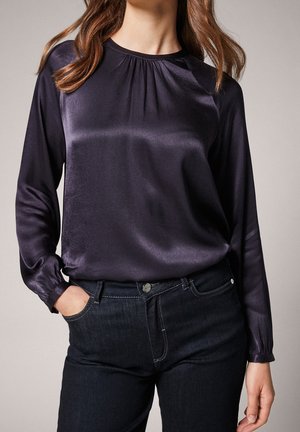 Kvinde iført en langærmet mørkelilla satinbluse stoppet ind i højtaljede sorte jeans, med den ene hånd i lommen.