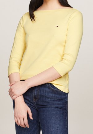 Femme portant un haut jaune clair à manches trois-quarts et un jean bleu foncé, les mains doucement croisées à hauteur de taille.