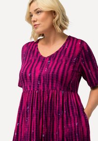 Sukienka z dekoltem w serek, wykonana z miękkiego materiału, z różowo-fioletowym wzorem i pionowymi liniami. Krótkie rękawy, zebra w talii oraz kieszenie.