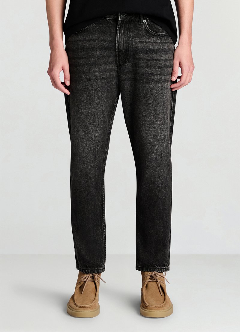 Jean en denim noir à coupe droite, présentant un léger délavage et une surface texturée. Associé à des chaussures marron clair, mettant en valeur des lacets et un style mocassin.