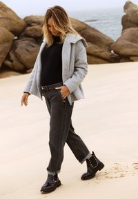 Manteau en laine gris avec capuche, chemise noire à manches longues, jean sombre avec les poignets retroussés et bottines noires à clous, marchant sur une plage.