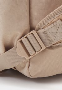 Béžová látková taška s plochým, texturovaným povrchem, nastavitelným popruhem a plastovou sponou s logem.