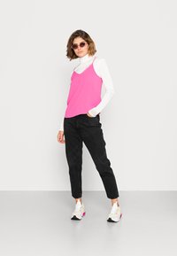 Camisola rosa com rendas em camadas sobre um suéter branco, combinada com jeans pretos ajustados e sapatilhas desportivas multicoloridas. Óculos de sol circulares.